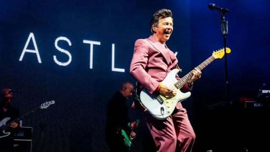 Rick Astley dressed in a pink suit on stage with a guitar