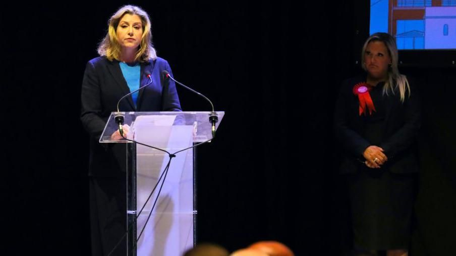 Penny Mordaunt giving a speech after being defeated by the Labour candidate standing on her right