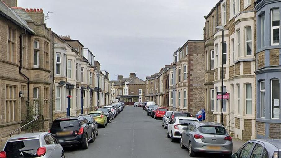 General street view of  Kensington Road in Morecambe, showing terraced houses on each side with bay windows, most of which have cars parked outside