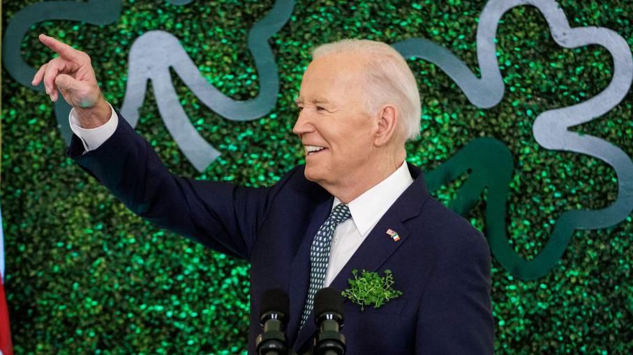 A man with white hair in front of a green background wearing a navy suit with white shirt and green tie, he is pointing