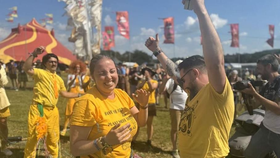 Mr Wilson and Ms Lawrence wearing yellow. They are celebrating and laughing with their hands in the air. Flags and festival tents are visible in the background and there are streamers all over them