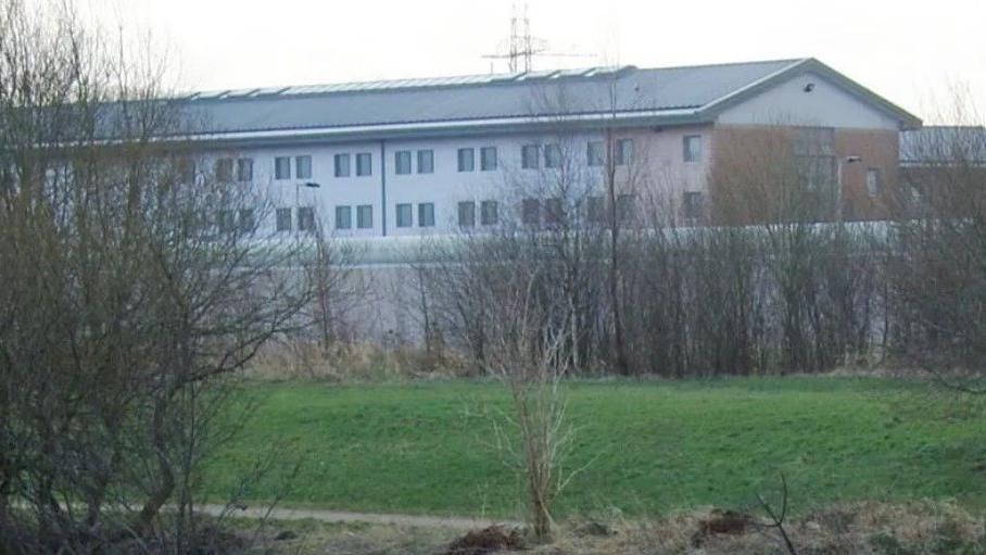 HMP Forest Bank, with grass and trees in the foreground of the picture