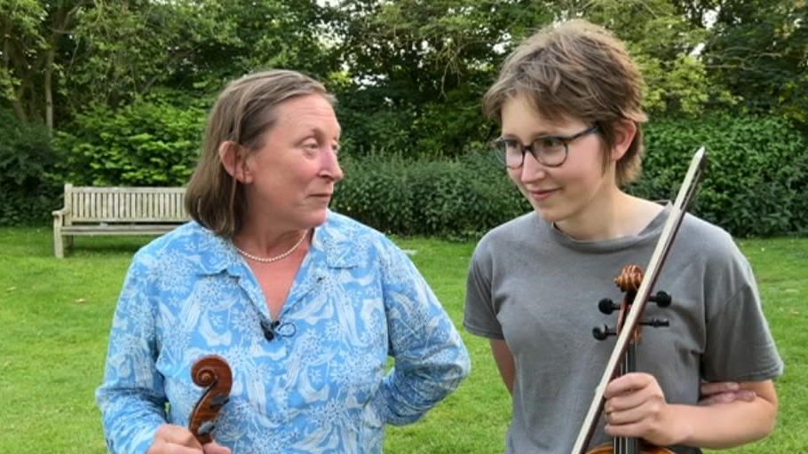 Anna wearing a blue shirt standing in a garden with her granddaughter Astrid Wilson who is wearing a grey shirt and holding a violin.