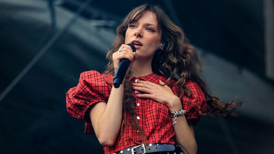 Holly Humberstone performing at a festival in August. Holly is a 22-year-old woman with long curly brown hair which she wears loose. She has blue eyes and holds a hand to her chest as she sings into a microphone. She's styled in a red tartan-look dress with puff sleeves and a studded black leather belt. 