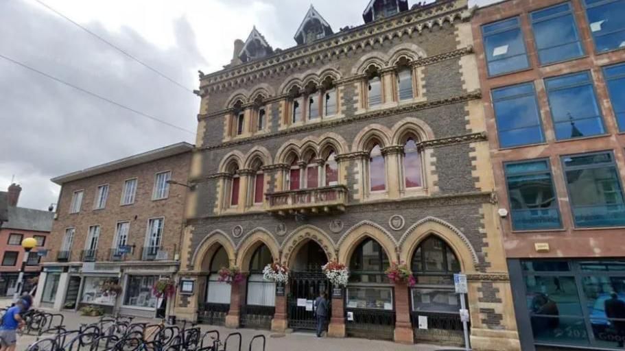 Hereford Museum and Art Gallery, a large brick building with several arches  on the ground floor, flanked by two other brick buildings. In front there are several metal stands for bicycles