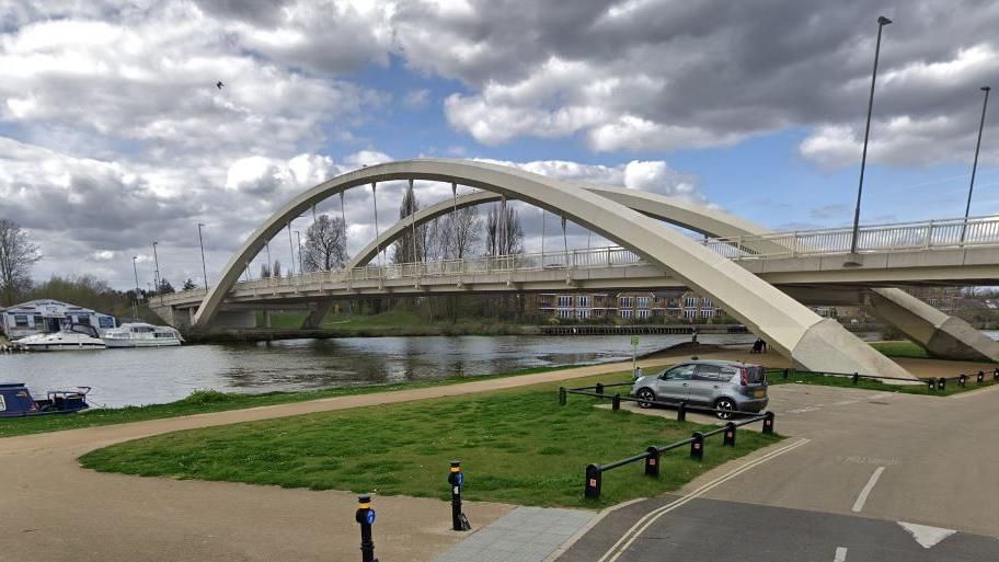 A large, modern bridge of two white arches is suspended over the river Thames with green verges, a carpark and boats nearby