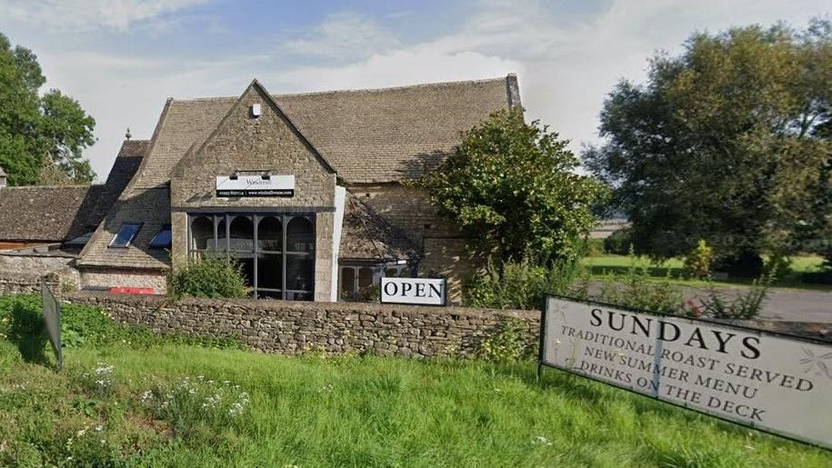 View of the pub from the road. It is a stone building with a sloping roof and a large floor-to-ceiling window at one end