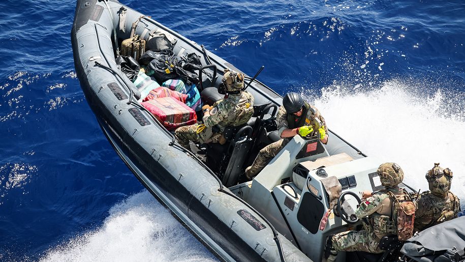 A rigid inflatable boat containing four personnel in camouflage outfits, seen from above. The boat appears to be cresting a wave in a choppy sea. 