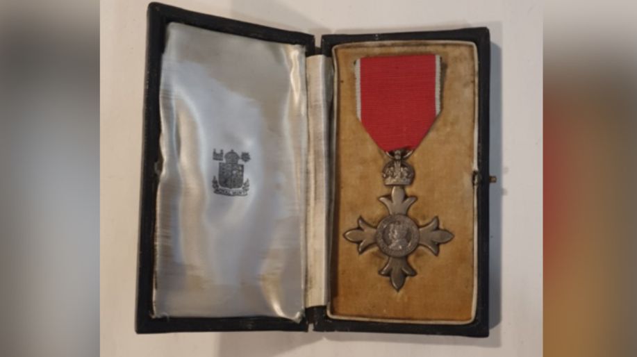 The MBE has a red ribbon and is in an old looking case and lies on a yellow background. The opposite side of the case is descorated with some silver material with some sort of insignia on it.
