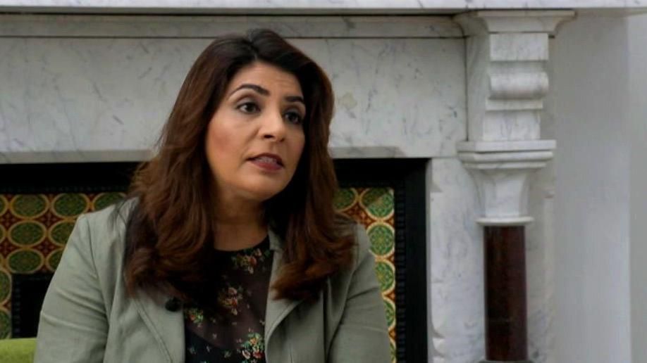 A woman with dark hair, wearing a grey suit top, sat in front of a white marble fireplace