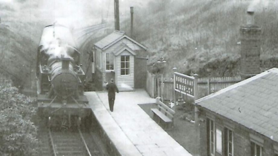 A black and white archive photo of a steam railway station with a train passing a signal box and a man walking on the platform