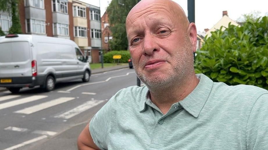 Steven Greatrex stands on the side of a road in front of a pedestrian crossing with a van going across it. He has a shaven head, stubble and is wearing a green and white polo shirt