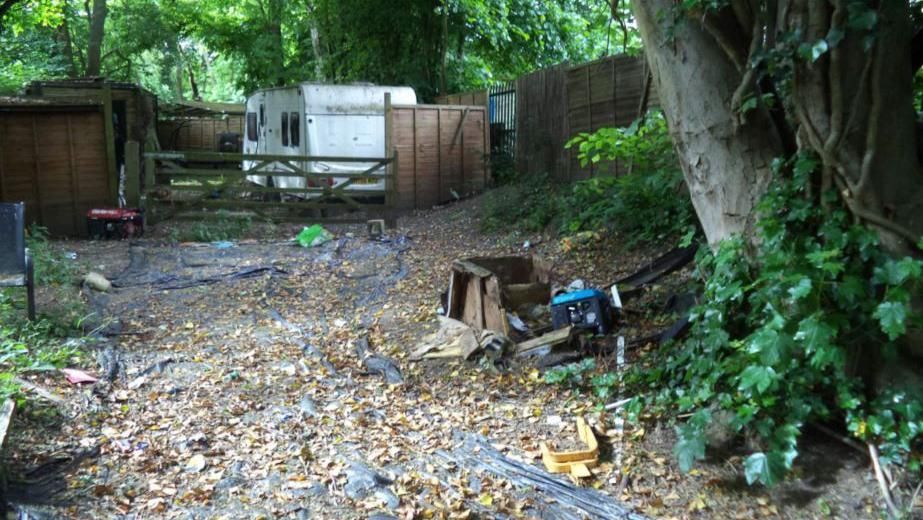 Illegal encampment on The Brambles in Salisbury. A caravan can be seen parked up behind a wooden gate with lots of other objects laid on the ground nearby. 