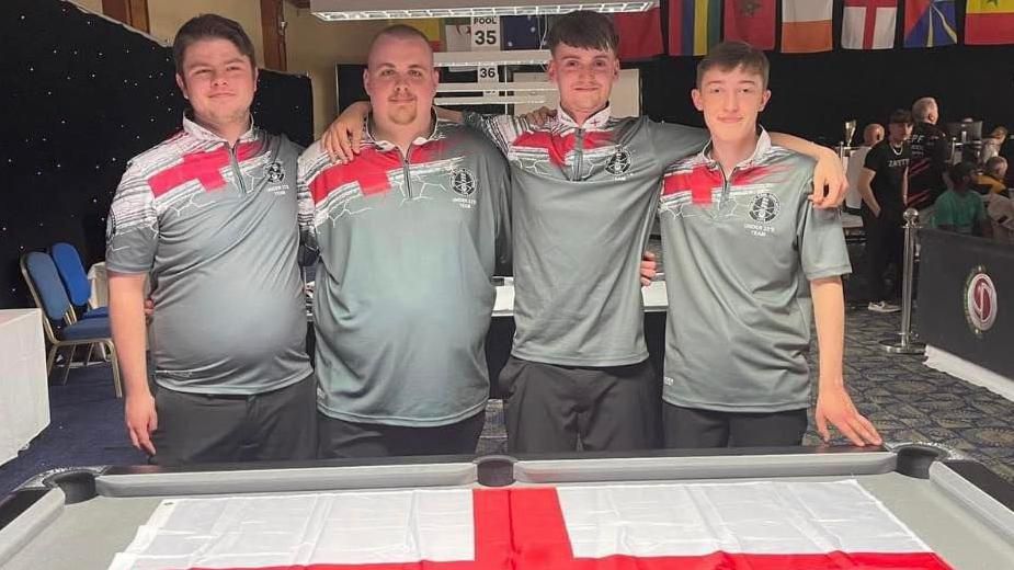 Four young male pool players stand behind a pool table draped with a St. George's flag. All wear grey T-shirts with a red cross at the top on the right shoulder.  They are, from left to right, Luke Cronin, Jake Cooper, Joshua Traynor and Matthew Ryall.