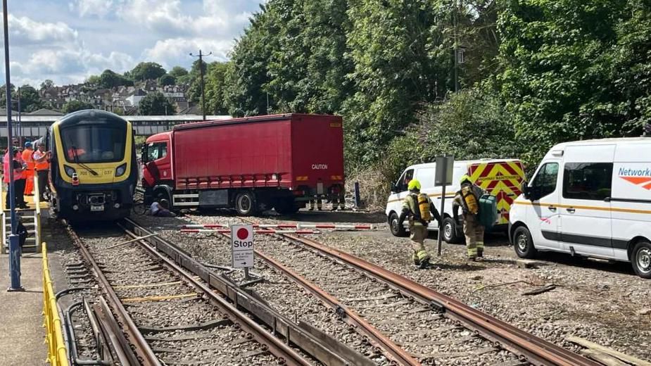 A train with a lorry parked next to it on train tracks. Network Rail vans and people in hi-vis jackets are on the scene