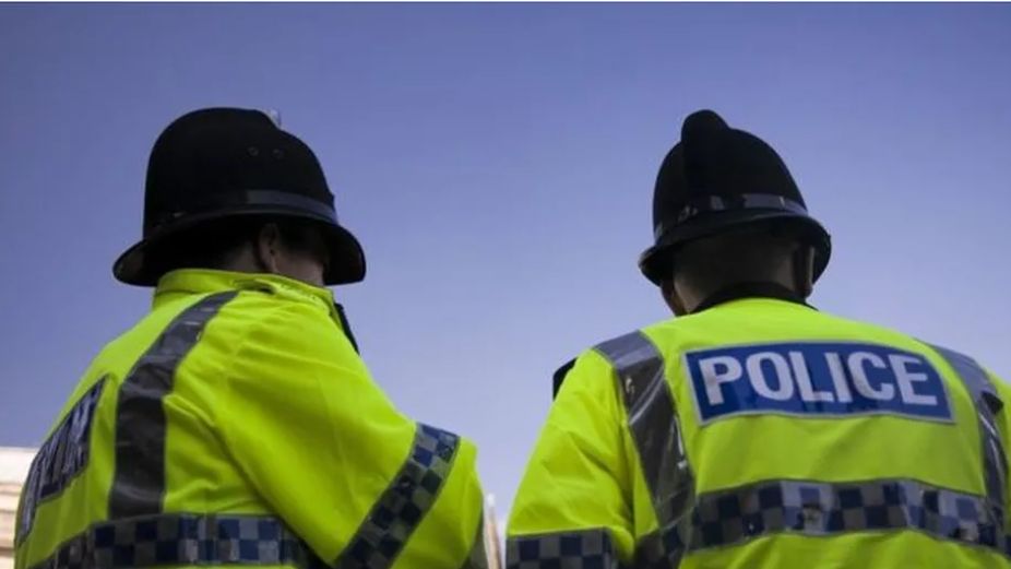 The back view of  two police officers wearing a lime green jacket with the word 'POLICE' in white writing on a blue background and black velvet police hats.