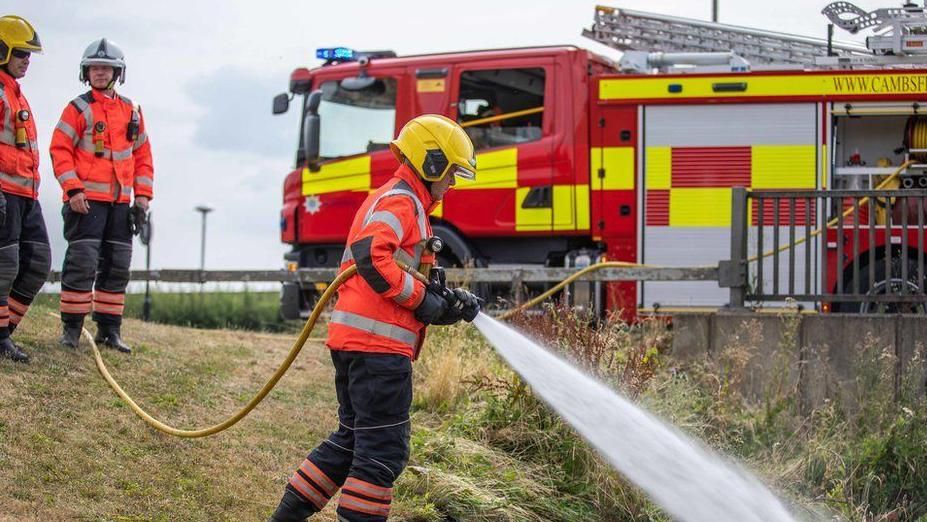 A firefighter hosing a grassfire with two other firefighters and a fire engine behind him