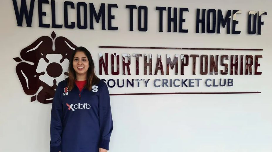 Nav Gahonia in a navy blue cricket tracksuit, standing next to a white wall with Northamptonshire cricket club branding on it.