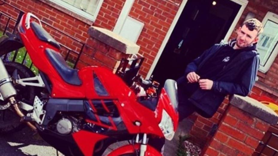 Brandern Holly standing outside a red brick house. He's wearing a blue rain jacket over a navy hoody and dark trousers. In front of him is a red motorbike and he is holding keys.