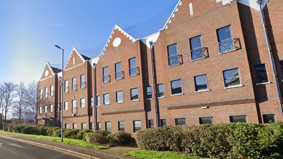 Goodman House, which is a large building made of bricks. It has many windows and is next to a main road, which has bushes to the side of it.