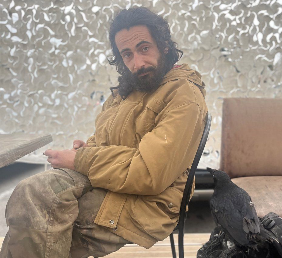 Julien, who lives in the mountains outside Quillan. He has long black hair and a black beard. He is wearing a beige coat. His pet crow sits next to him.