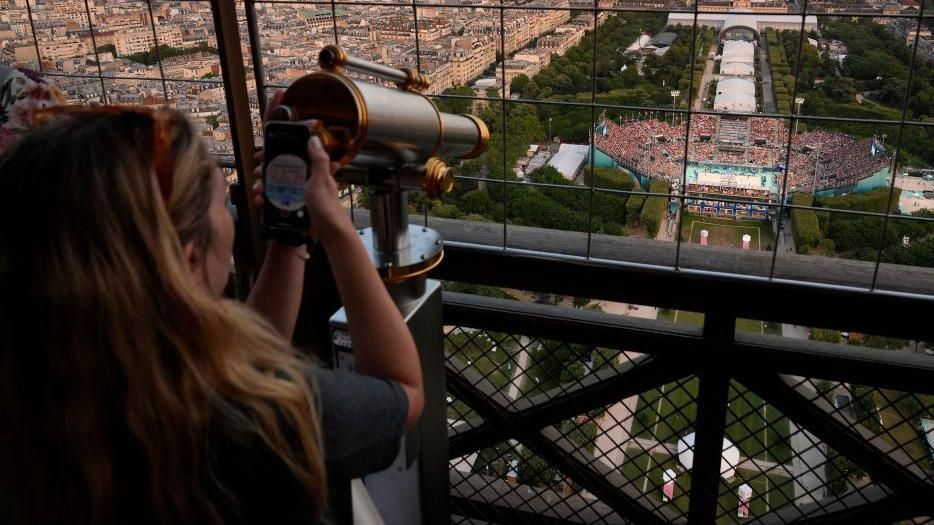 A woman uses a telescope on the Eiffel Tower to watch a volleyball match in a stadium below