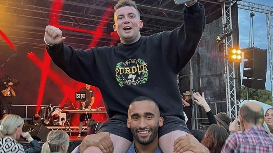 Man carries another man on his shoulders with their backs to a festival stage