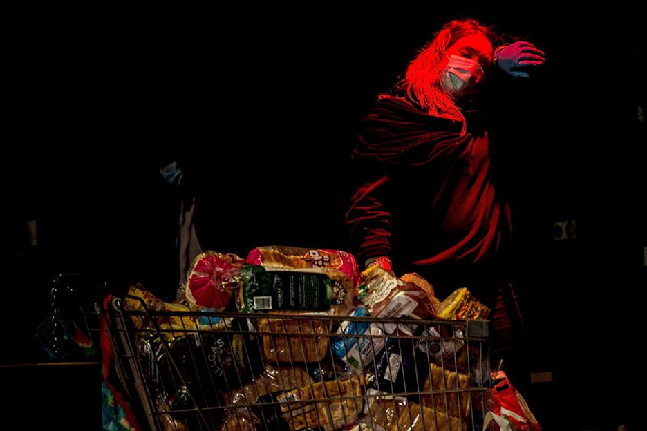 A woman wearing a face mask stands next to a shopping trolley full of food