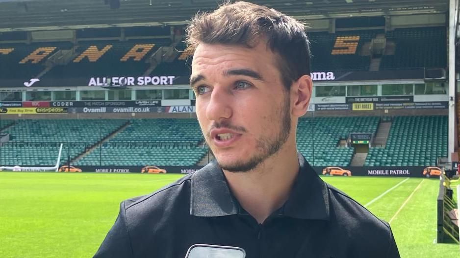 Norwich City midfielder Liam Gibbs speaking pitchside by an empty Carrow Road