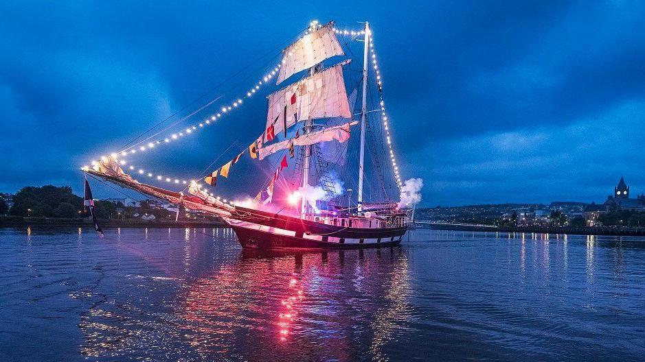 An illuminated tall ship sailing down the foyle at dusk, the city's Guildhall is seen in the background