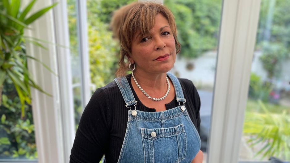 Sam Van Der Heijden. She has brown hair which is tied up, and is wearing a pearl necklace and hoop earrings. She standing in a house, with a garden seen behind her. She is wearing blue denim overalls and a black top. 