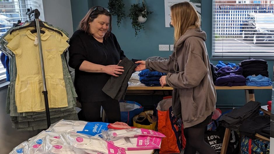 A woman in a black t-shirt is holding some grey children's trousers. A woman to her right, wearing a brown hoodie, is passing the trousers to her. To the left is a clothes rail with coloured checkerboard school dresses hanging on it. There are also tables piled with other clothing.