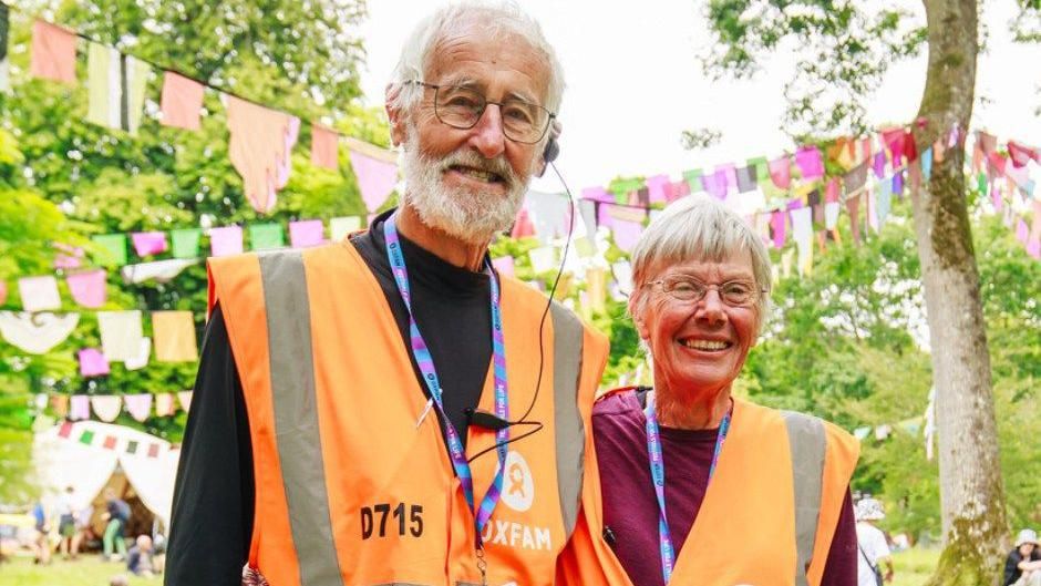 Graham and Judy Cole wearing orange Oxfam hi-vis vests and lanyards. They are standing amongst some trees that have bunting hanging between the branches