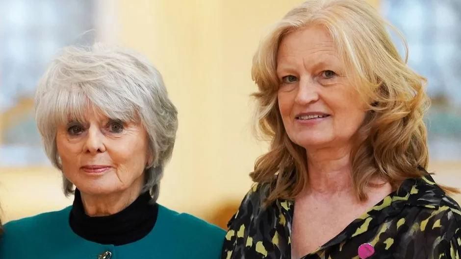 Diana Parkes stands to the left of the frame. She has short, grey hair and wears a black turtle neck jumper, with a teal blue collarless jacket with gold detailed buttons. She looks directly at the camera and is standing next to her friend, Hettie Barkworth-Nanton, who has mid-length, wavy, ginger hair and wears a V-neck yellow and black abstract printed blouse