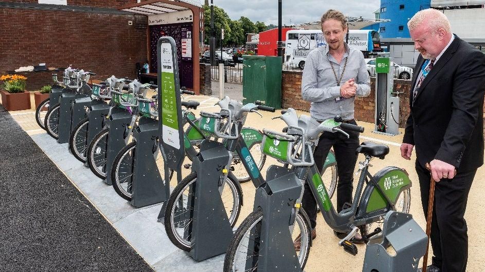 Two men stand near a row of locked bikes for hire. One man is wearing a grey shirt and black trousers, the other is wearing a black suit and leaning on a cane. Behind them a bus and some parked cars are visible