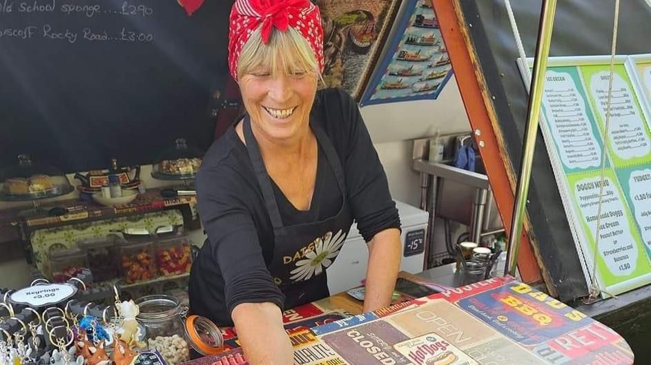 A woman wearing red bandana and a black apron with a picture of a daisy on it, leans forward over the counter of a stall selling keyrings and other items