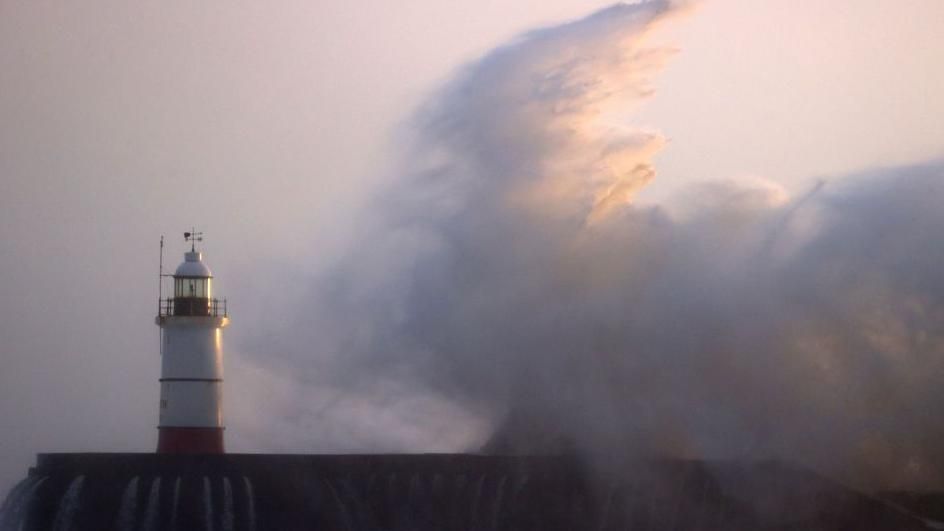 Photograph of a lighthouse with large waves crashing in the distance and onto a pier