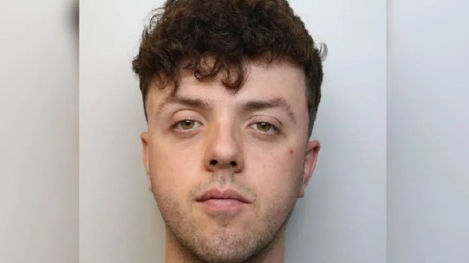 A police mugshot of Sam Powers. He has dark brown hair, short at the sides and longer on top. 