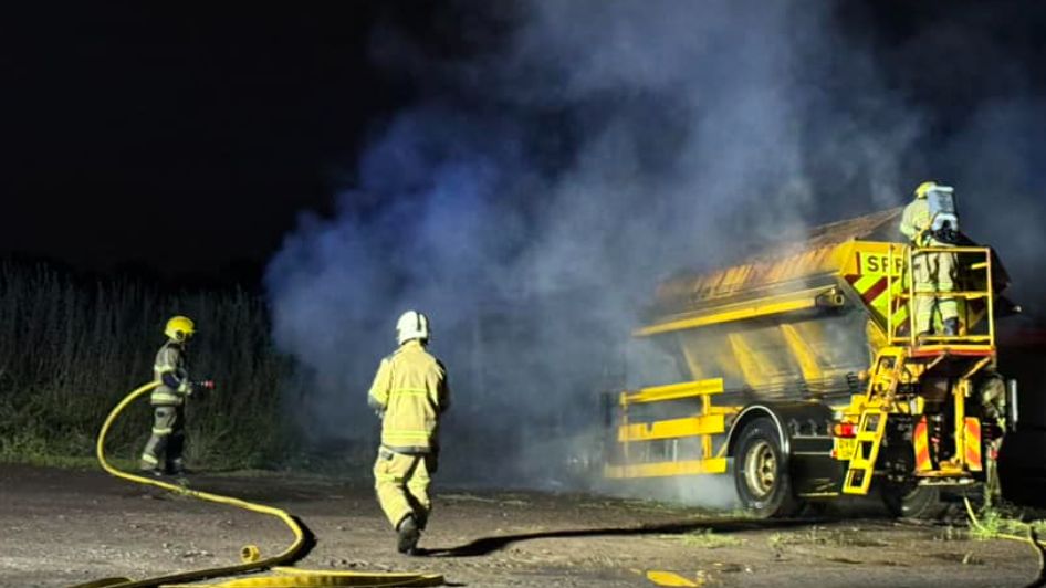 Smoke surrounds a yellow gritting lorry under dark skies, with firefighters to the back and side of the vehicle with hoses