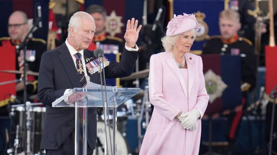 King Charles waves to the crowds in front of a podium beside Queen Camilla