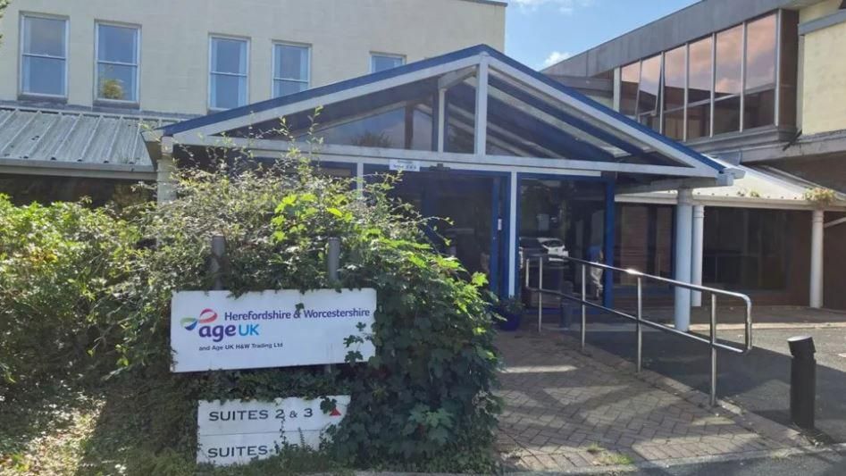 Age UK Herefordshire & Worcestershire building with the sign outside, amid bushes, outside a glass awned porch