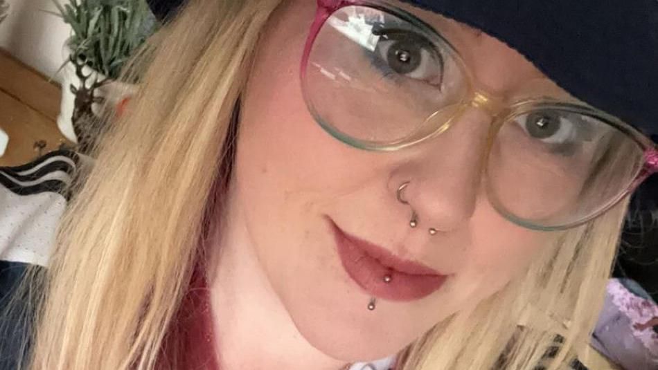 Tamara Beattie. Tamara is a young woman with long blonde hair worn loose under a navy bucket hat. She has her nose and lip pierced and wears rainbow coloured glasses.