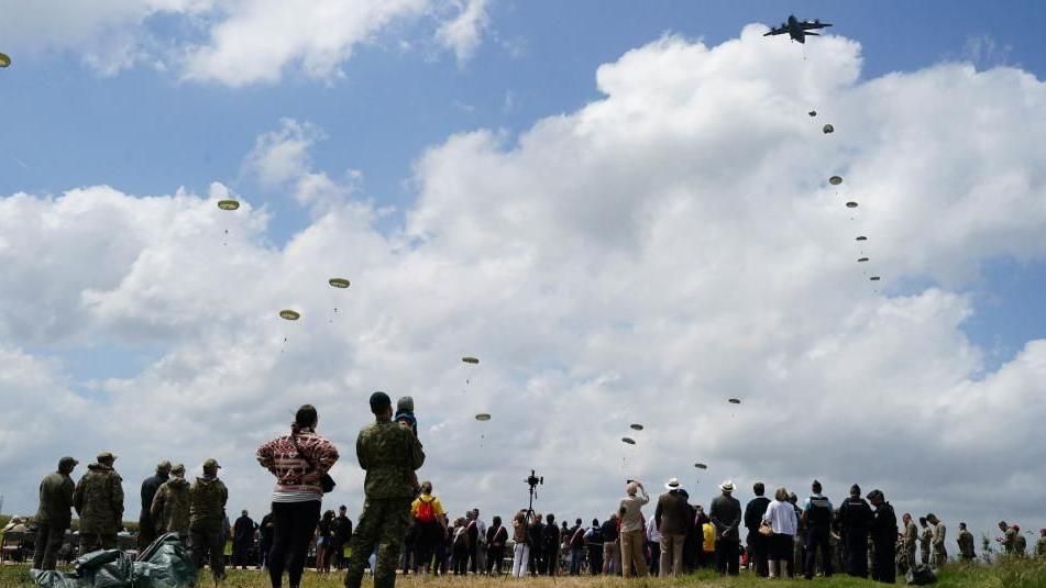 Crowds stand to watch parachutes drift to the ground
