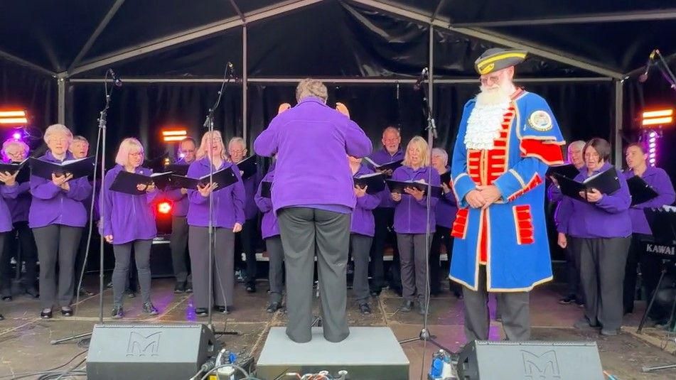 Martin Wood, wearing his town crier regalia, listens at the front of an outdoor stage while the choir, dressed in purple fleeces, perform the song