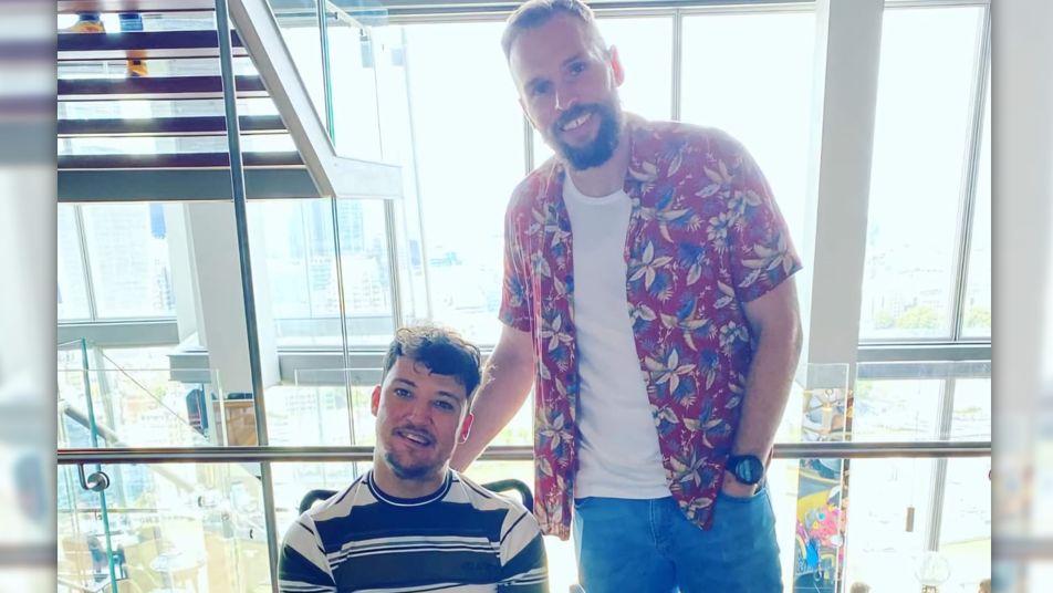 Two men smile at the camera, one is in a wheelchair