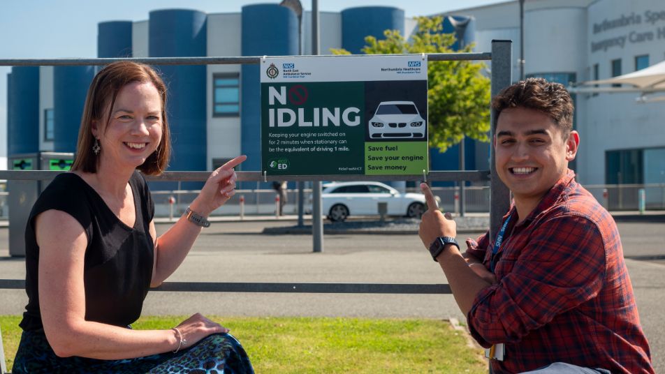 Two people pointing at a sign which reads: "No Idling. Keeping your engine switched on for 2 minutes when stationary can be equivalent to driving 1 mile"