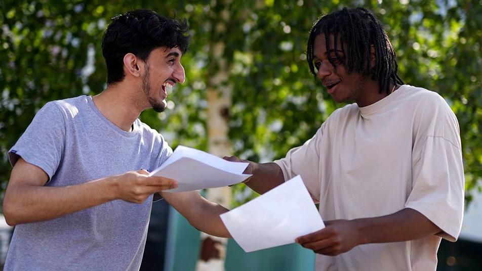 Two students from Ark Globe Academy in London receive their results. One wears a grey T-shirt and is holding his results in his hand and laughing, while his classmate, wearing a beige T-shirt, points at the paper.