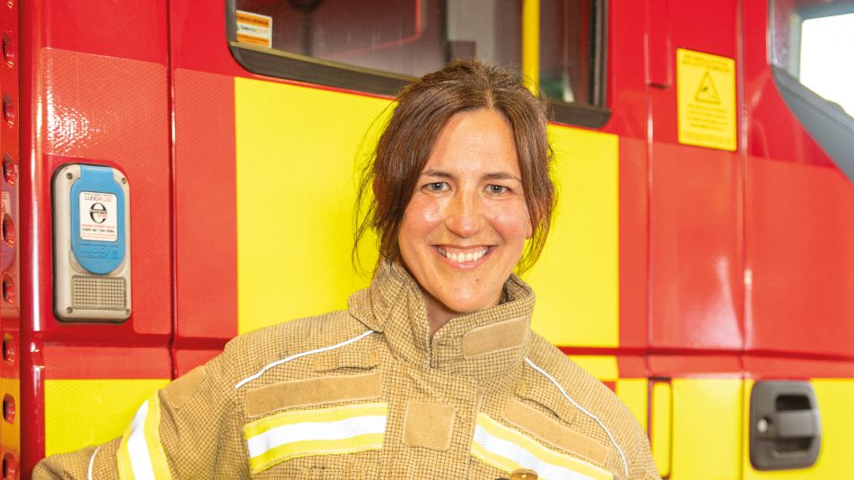 A female firefighter stands in front of a red and yellow fire engine in a firefighters jacket