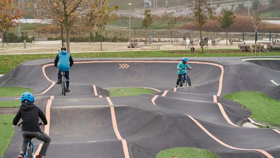 Two children and a man cycle on a black-surfaced track in a park which has slopes, bends and curves. The youngsters are wearing blue cycle helmets and are riding BMX bikes. 