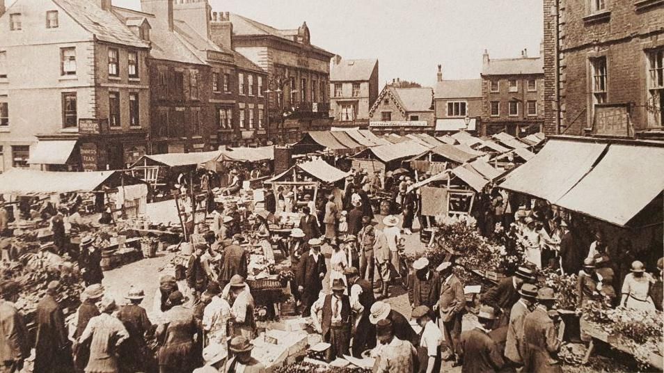 A black and white photo showing lots of people in the 1920s around many stalls in a market place